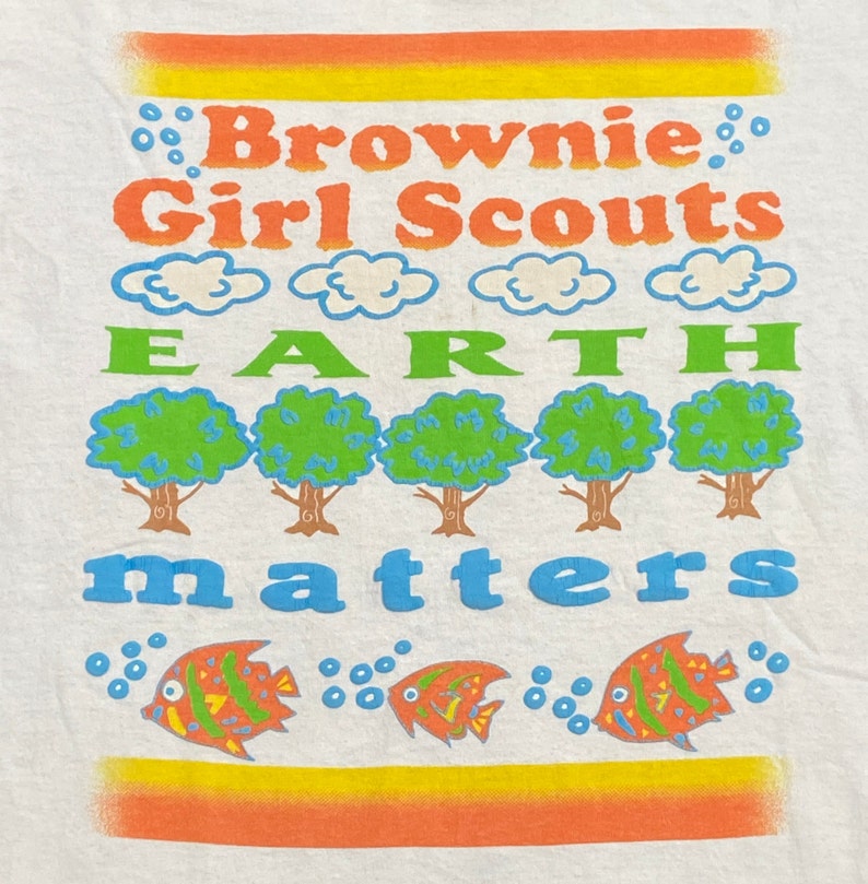 K&ouml;nnte beinhalten: Wei&szlig;es T-Shirt mit einem bunten Grafikdesign. Das Design zeigt den Text "Brownie Girl Scouts" und "Earth Matters" mit einer Reihe von B&auml;umen und Fischen.