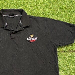 May include: Black polo shirt with a collar and short sleeves. The Guinness logo is embroidered on the chest. The logo features a harp and the Guinness name in white and red.