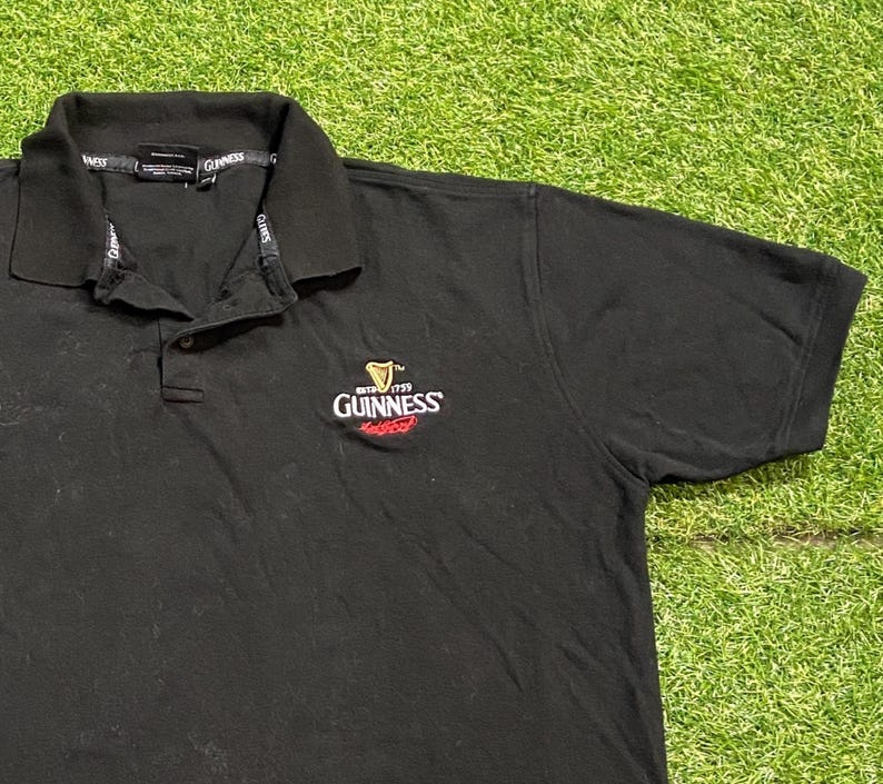 May include: Black polo shirt with a collar and short sleeves. The Guinness logo is embroidered on the chest. The logo features a harp and the Guinness name in white and red.