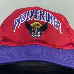 Puede incluir: Gorra de béisbol roja y morada con un logotipo bordado en blanco y morado que dice "Wolverine!" con una imagen de dibujos animados de la cara de Wolverine en el centro.