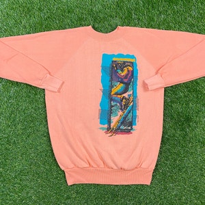 May include: A peach-coloured sweatshirt with a graphic print of a person skiing down a mountain. The graphic is in a blue, green, yellow, and red colour scheme.