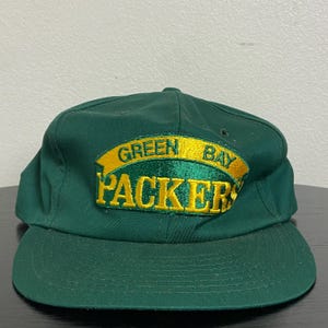 Puede incluir: Gorra de béisbol verde con visera curva. La gorra presenta las palabras "GREEN BAY PACKERS" bordadas en oro y verde. La gorra está hecha de una tela duradera y tiene un diseño clásico y ajustable.