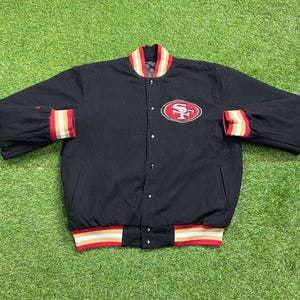 May include: Black button-up jacket with red, gold, and white striped accents on the collar and cuffs. Features a red and gold embroidered logo on the chest. The jacket is lying on a green surface.