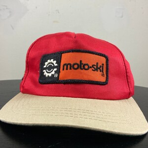 Peut inclure: Casquette de baseball rouge et beige avec un patch noir et orange qui dit "moto-ski" avec un logo d'engrenage.
