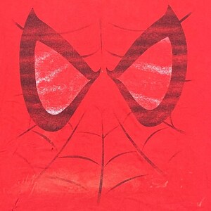 Puede incluir: Camiseta roja con un gr&aacute;fico de m&aacute;scara de Spider-Man en blanco y negro. La m&aacute;scara tiene un dise&ntilde;o de telara&ntilde;a y est&aacute; centrada en la camiseta.