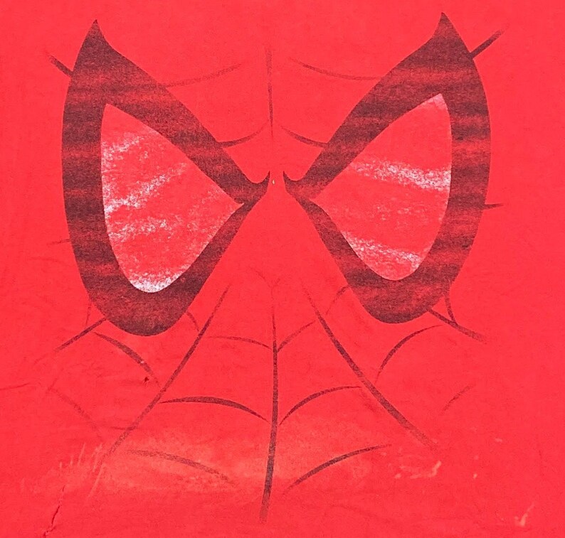 Puede incluir: Camiseta roja con un gr&aacute;fico de m&aacute;scara de Spider-Man en blanco y negro. La m&aacute;scara tiene un dise&ntilde;o de telara&ntilde;a y est&aacute; centrada en la camiseta.