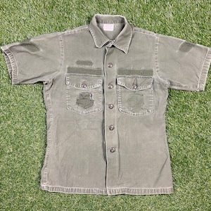 以下が含まれることがあります： 2つの大きなポケットが付いた緑色の半袖ボタンダウンシャツ。色あせている。