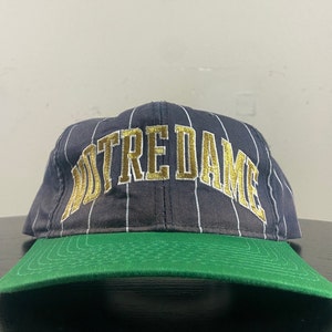 May include: A black and white striped baseball cap with a gold embroidered "NOTRE DAME" logo. The cap has a green underside.