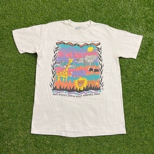 May include: White t-shirt featuring a colorful graphic of zoo animals, including a giraffe, zebra, elephant, and lion. The text "SAN DIEGO ZOO WILD ANIMAL PARK" is printed below the graphic. The shirt is made of a soft material.