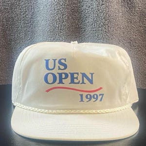 Op de afbeelding: Crèmekleurige baseballcap met de tekst "US OPEN" in blauw, een rode gebogen lijn en het jaartal "1997" in blauw. De cap heeft een wit koorddetail rond de rand.