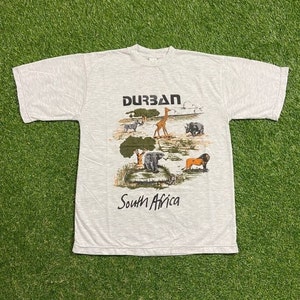 May include: Light gray t-shirt featuring a graphic of African wildlife, including a giraffe, zebra, and lion. The word "Durban" is printed above the image, and "South Africa" is printed below. The shirt is laid out on a green surface.