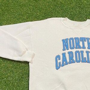 May include: Cream-colored sweatshirt with the words "NORTH CAROLINA" in light blue arched lettering. The long-sleeved top is made of a soft material and is suitable for casual wear.