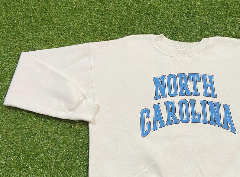 May include: Cream-colored sweatshirt with the words "NORTH CAROLINA" in light blue arched lettering. The long-sleeved top is made of a soft material and is suitable for casual wear.