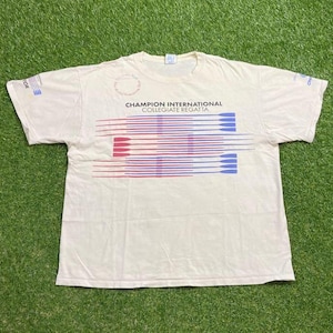 Puede incluir: Camiseta color crema con el texto "Champion International Collegiate Regatta" en negro y un diseño de líneas horizontales rojas y azules. La camiseta tiene mangas cortas y cuello redondo.