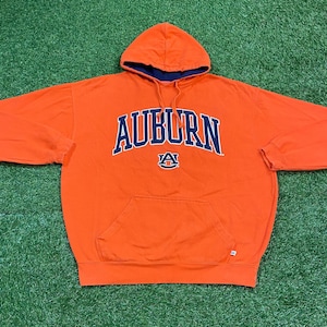 May include: An orange hooded sweatshirt with the word "AUBURN" in navy blue arched across the chest. Below the word "AUBURN" is a navy blue "A" logo. The hood has a navy blue lining, and the sweatshirt has a front pocket.