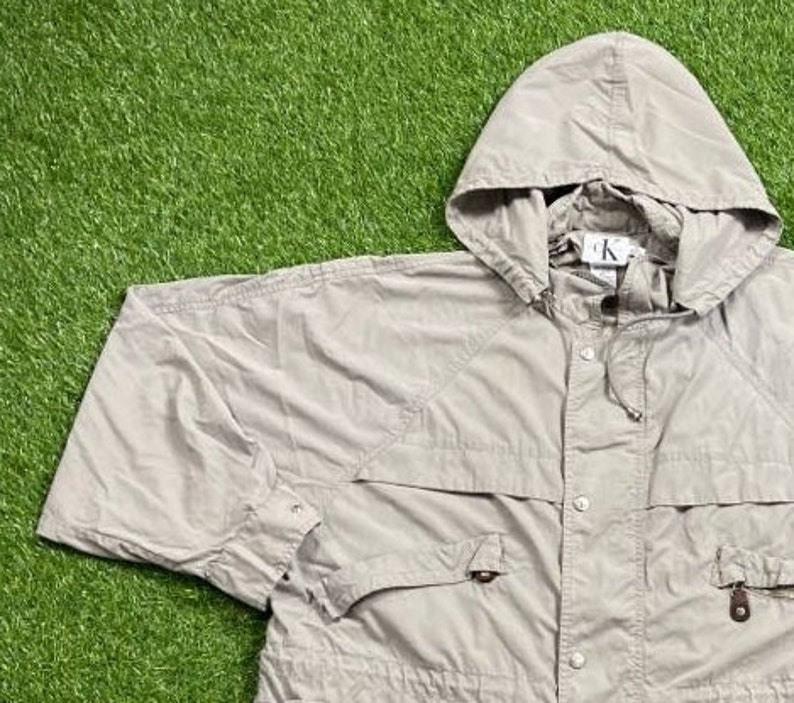 Pu&ograve; includere: Giacca con cappuccio color beige chiaro con coulisse e chiusura con bottoni. La giacca ha due tasche frontali e un piccolo logo sul cappuccio. La giacca &egrave; realizzata in materiale leggero.