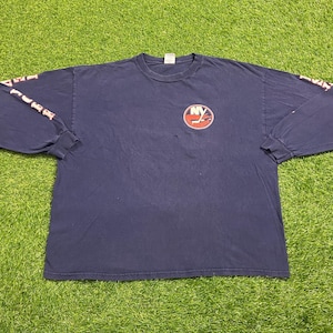 May include: Navy blue long-sleeve t-shirt featuring the New York Islanders logo on the chest. The sleeves have the team name printed in red. The shirt is laid out on a green grassy surface.