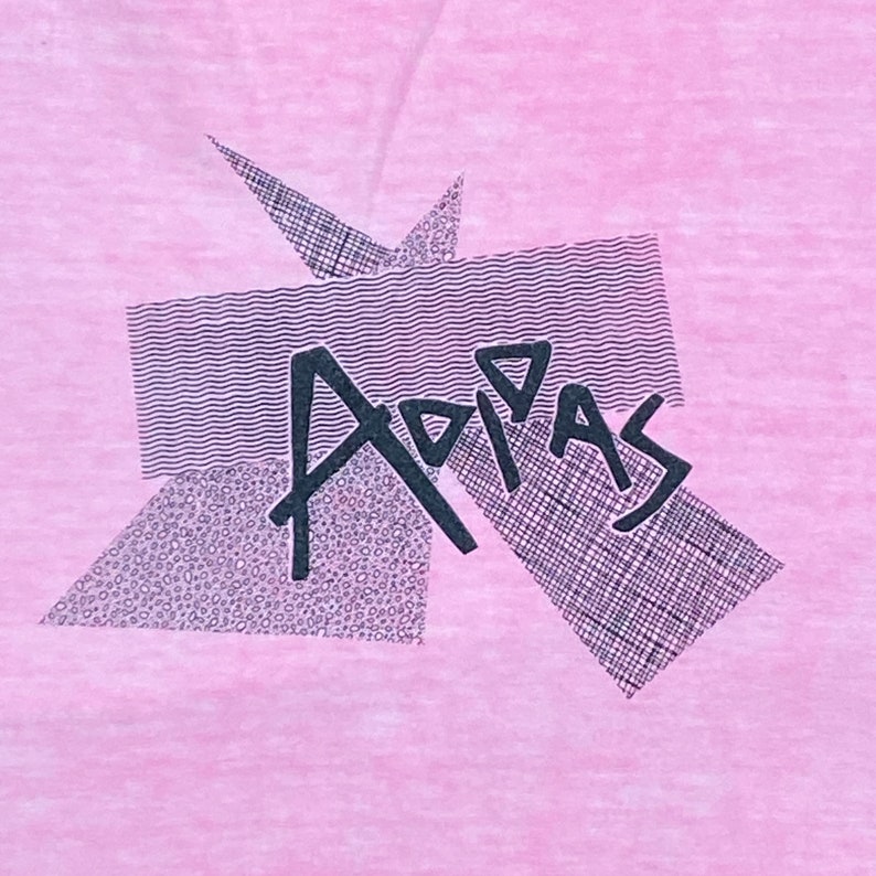 Peut inclure: Un t-shirt rose avec un motif graphique noir du mot "Adidas" dans un design g&eacute;om&eacute;trique.