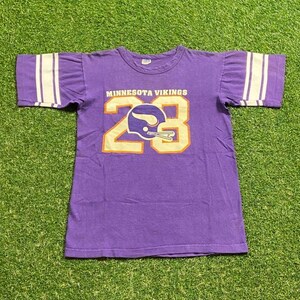 May include: Purple vintage t-shirt with the text "MINNESOTA VIKINGS" above the number "23" in gold. The number features a Viking helmet graphic. White stripes adorn the sleeves.