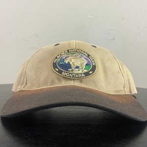 May include: A tan baseball cap with a brown brim. The front of the cap features a patch with the words "Glacier National Park" and a picture of a bear. The words "Montana" are printed below the bear.