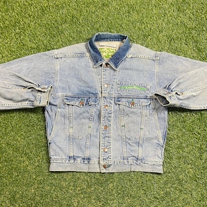 May include: A light blue denim jacket with a green and white logo that says "Cool Blue" on the left chest. The jacket has a button-up front and two pockets on the front.