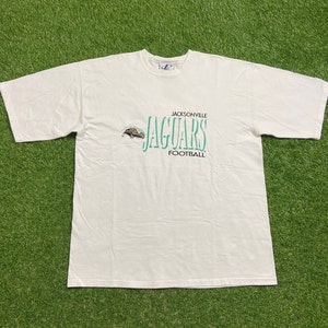 May include: A white t-shirt featuring the text "JACKSONVILLE JAGUARS FOOTBALL" in teal. The shirt has a small logo of a jaguar. The shirt is laid flat on a green surface.