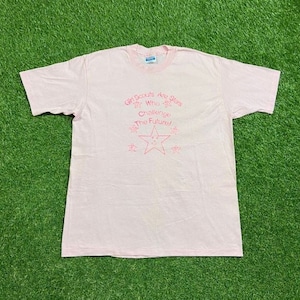 Puede incluir: Una camiseta rosa claro con cuello redondo y mangas cortas. La camiseta presenta el texto "Girl Scouts Are Stars Who Challenge The Future!" en rosa, junto con gr&aacute;ficos de estrellas. La camiseta est&aacute; tendida sobre una superficie verde.