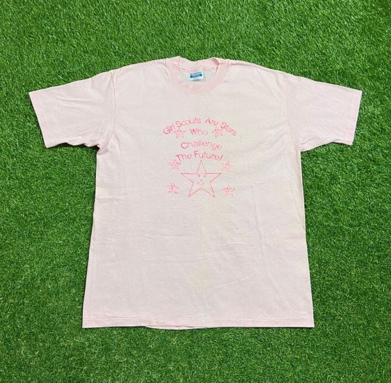 Puede incluir: Una camiseta rosa claro con cuello redondo y mangas cortas. La camiseta presenta el texto "Girl Scouts Are Stars Who Challenge The Future!" en rosa, junto con gr&aacute;ficos de estrellas. La camiseta est&aacute; tendida sobre una superficie verde.