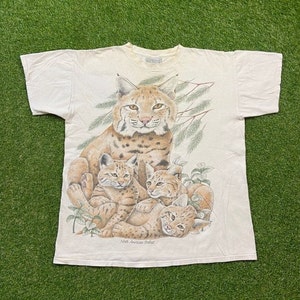 May include: A white t-shirt with a graphic of a North American Bobcat and three kittens. The bobcat is brown and black with spots. The kittens are smaller and have similar markings. The graphic is surrounded by green foliage.