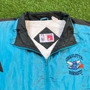 Może przedstawiać: Niebieska i czarna kurtka wiatr&oacute;wka z logo Charlotte Hornets i haftowanym napisem "Charlotte Hornets" z przodu. Kurtka zapinana na zamek błyskawiczny, z białą podszewką. Kurtka ma metkę marki "Color" z przodu.