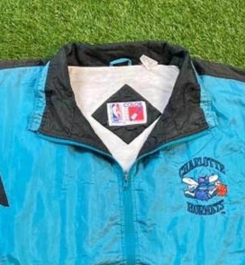 Może przedstawiać: Niebieska i czarna kurtka wiatr&oacute;wka z logo Charlotte Hornets i haftowanym napisem "Charlotte Hornets" z przodu. Kurtka zapinana na zamek błyskawiczny, z białą podszewką. Kurtka ma metkę marki "Color" z przodu.
