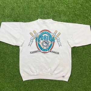May include: White sweatshirt with a vintage Florida Marlins baseball logo. The logo features a marlin fish, two crossed bats, and the text "Baseball Florida Club National League".