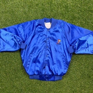 Pode incluir: Uma jaqueta bomber de cetim azul com um personagem de desenho animado vermelho, laranja e amarelo bordado no peito esquerdo. A jaqueta tem um fecho de correr e um colarinho, punhos e bainha canelados.