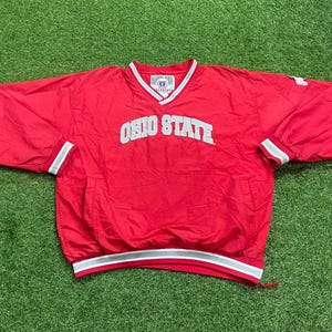 May include: A red long-sleeve pullover windbreaker jacket featuring the text OHIO STATE in white lettering across the chest. The garment has a V-neck collar with white and grey trim, matching striped cuffs, and a drawstring hem at the bottom.
