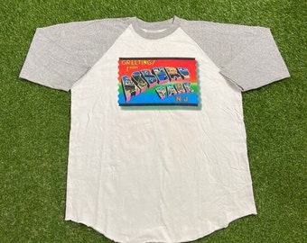May include: A white and grey baseball t-shirt with a colorful "Greetings from Asbury Park, N.J." postcard graphic on the front.