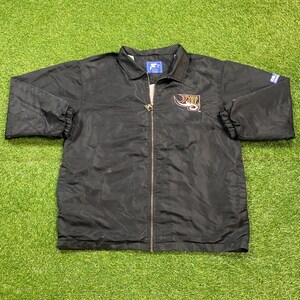 Peut inclure: Veste noire Starter avec fermeture éclair intégrale. La veste présente un logo sur la poitrine et un logo Starter sur la manche. La veste est posée sur une surface verte.