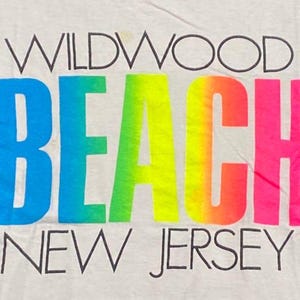 May include: A close-up of a white t-shirt with the words "WILDWOOD BEACH NEW JERSEY" printed on it. The word "BEACH" is in a gradient of blue, green, yellow, orange, and pink.