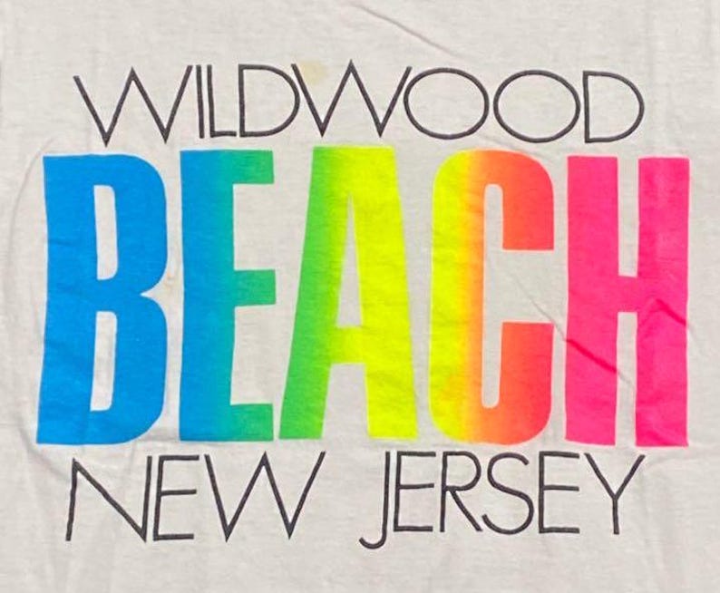 May include: A close-up of a white t-shirt with the words "WILDWOOD BEACH NEW JERSEY" printed on it. The word "BEACH" is in a gradient of blue, green, yellow, orange, and pink.