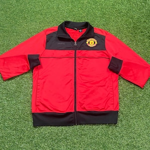 May include: A red and black zip-up track jacket with the Manchester United logo on the chest. The jacket has a black collar, cuffs, and hem, with the MUFC logo on the chest.