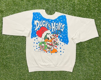 Può includere: Felpa bianca con una grafica blu e bianca di un'anatra di cartone animato che indossa un cappello da Babbo Natale e tiene un bastone di zucchero. Il testo "Duck the Halls" è stampato sulla grafica.