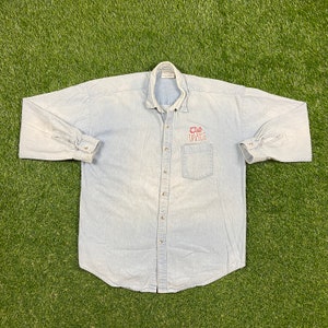 May include: Light blue denim button-down shirt with long sleeves. Features a chest pocket and the embroidered text "Club Dance" in red. The shirt is laid flat on a green surface.