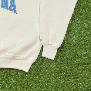 May include: White crew neck sweatshirt with blue text. The sweatshirt has a ribbed cuff and a relaxed fit. The garment is made of a soft material and is suitable for casual wear. The sleeve cuff has a small logo.