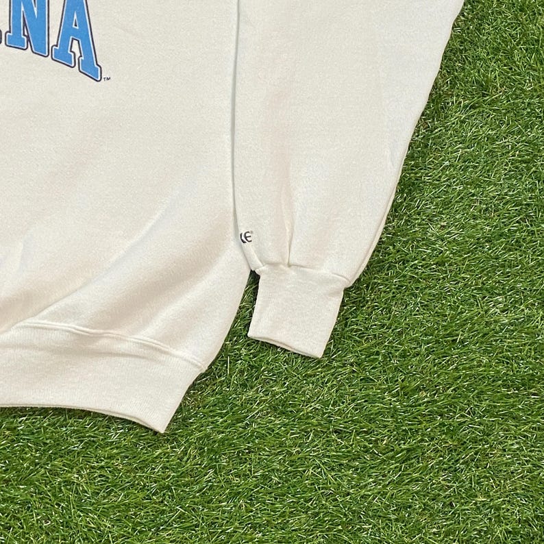 May include: White crew neck sweatshirt with blue text. The sweatshirt has a ribbed cuff and a relaxed fit. The garment is made of a soft material and is suitable for casual wear. The sleeve cuff has a small logo.