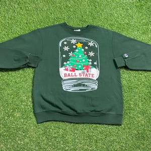 May include: A dark green sweatshirt featuring a snow globe design with a Christmas tree, snowflakes, and the text "BALL STATE". The tree is decorated with ornaments and topped with a yellow star. The sweatshirt has long sleeves and a Champion logo on the cuff.