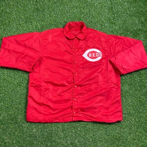 Puede incluir: Una chaqueta de nylon roja con cierre de botones blancos y un logotipo "REDS" blanco en el pecho.