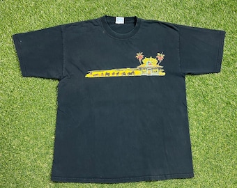 May include: Black t-shirt with a graphic featuring a tropical scene with palm trees and a building, and the text "Cafe Del Sol". Below the scene is a yellow horizontal line with motorcycle silhouettes. The shirt is laid out on a green surface.