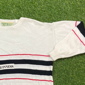 May include: White sweater with red and black horizontal stripes and a black patch with the word "GUINNESS" in white letters. The sweater has a red and black striped cuff on the right sleeve.