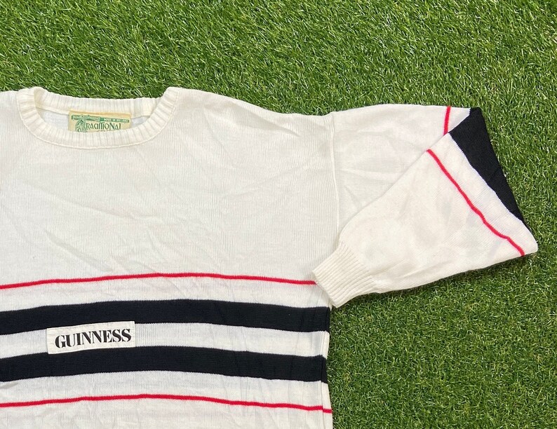 May include: White sweater with red and black horizontal stripes and a black patch with the word "GUINNESS" in white letters. The sweater has a red and black striped cuff on the right sleeve.