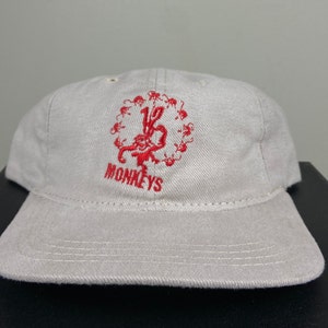 May include: A beige baseball cap with a red embroidered logo of 12 monkeys surrounding a monkey with the word "MONKEYS" below.