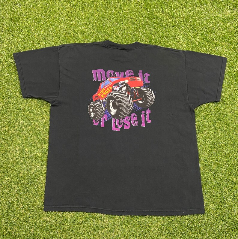 Może przedstawiać: Czarny T-shirt z grafiką czerwonego monster trucka z napisem "Move it or Lose it" nadrukowanym na fioletowo.